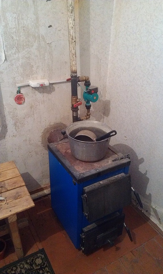 A photo: Tucked in a corner next to a folded-up wooden stool sits a small, dusty old-fashioned water boiler, in bright blue and black, with two under-stove compartments, connected to a series of worn pipes and behind it. A metal water container sits on the surface with a smaller metal household water scoop with a black handle inside it.