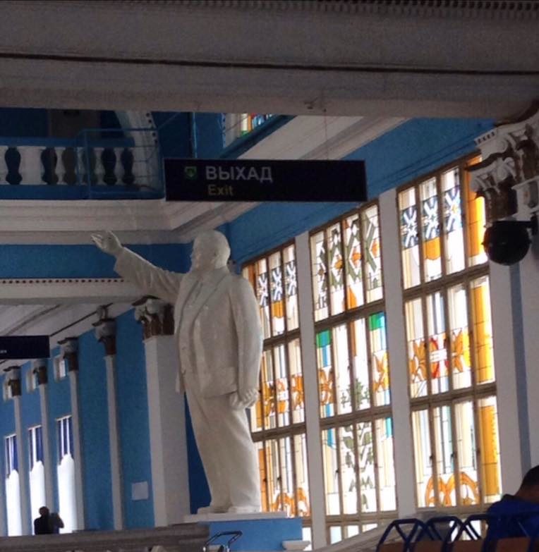 Photo of a white statue of Vladimir Lenin with arm raised, saluting, located indoors in front of stained glass windows and blue and white walls.