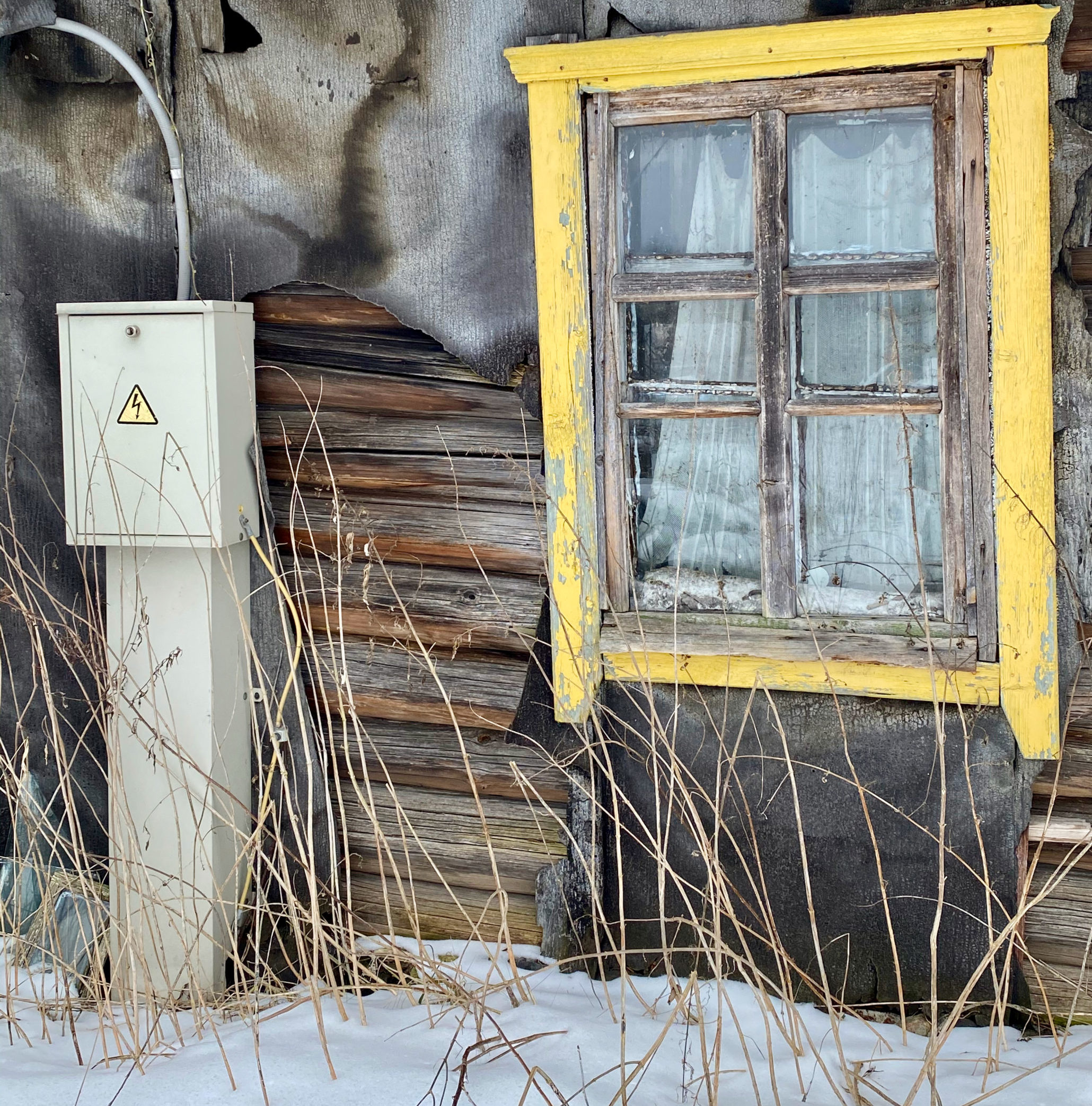 Side of abandoned wooden building with a bright yellow window frame and an electricity box.