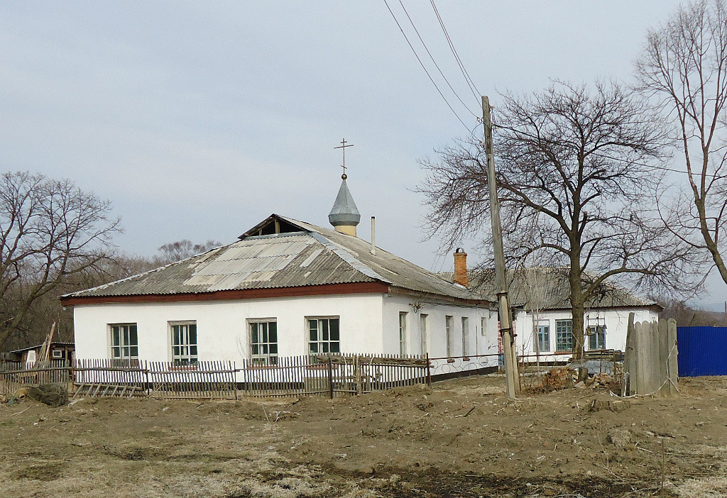 A photo of a low, white-washed building with a metal pitched roof. A small orange chimney and a simple onion dome sit on top. The church is surrounded by a low, wooden stick fence and brown dirt ground. To the right is a telephone pole with wires/cables extending from it.
