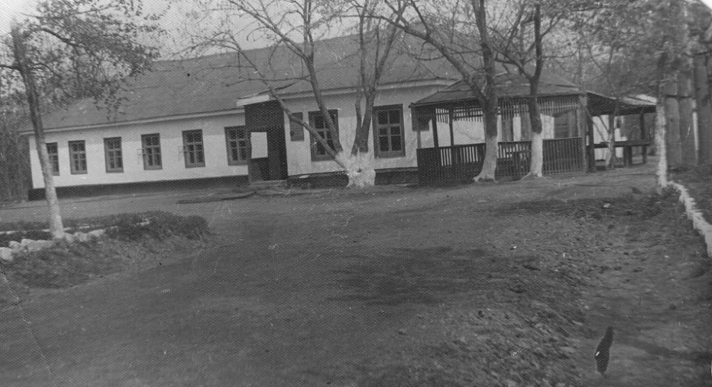 A black and white photo showing a long, white one-storey building with a pitched roof and a row of rectangular framed windows, with a sheltered outdoor area to the right. A few bare trees stand in front of the building, with their bottom sections painted white.