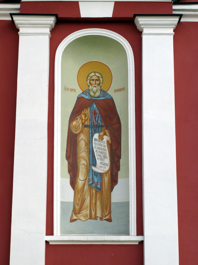 A photo of an icon of a male saint with an orange-gold halo, clad in orange-gold vestments with a red cape and blue-and-red over vestment. The saint holds an open scroll in their left hand and his right hand is held in the gesture blessing. The icon is set between two white pillars against a red wall.
