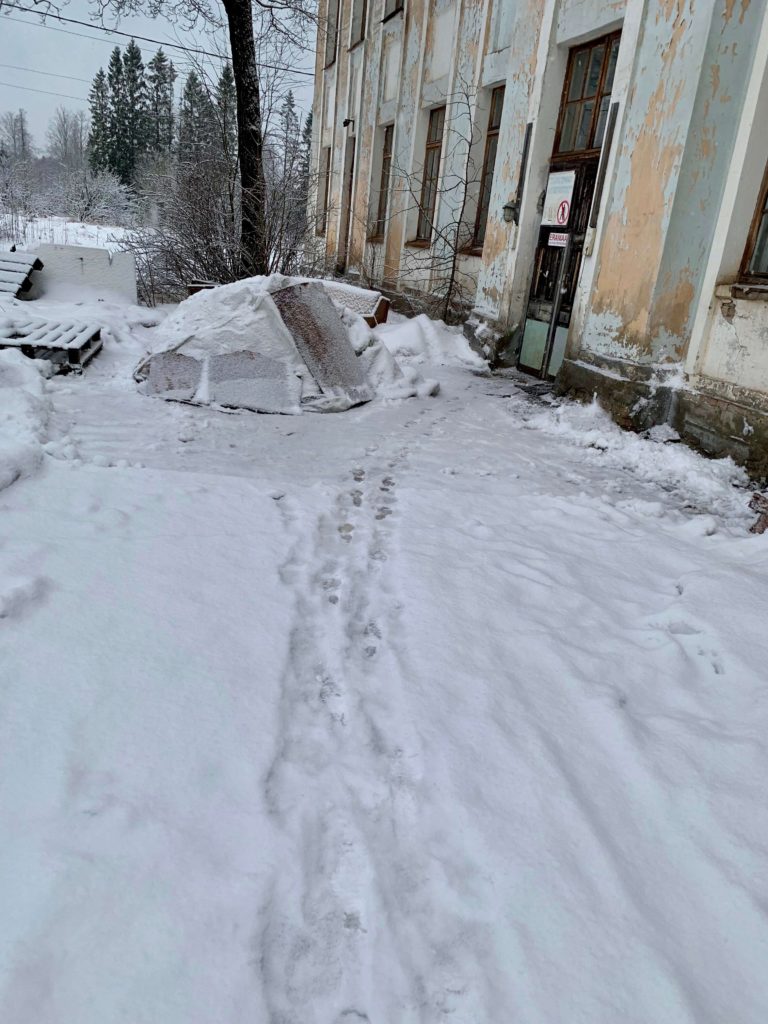 Photo of the side of a large and weathered building, this one has many columns up the side of the building and peeling light blue paint. A sign on the door displays an outline of a man encircled and barred with red: no entry, private. Immediately in front of the door are snow-covered piles of scrap material and trees growing close to the walls. In the background are bigger trees.
A trail of footprints in the snow lead to the door.