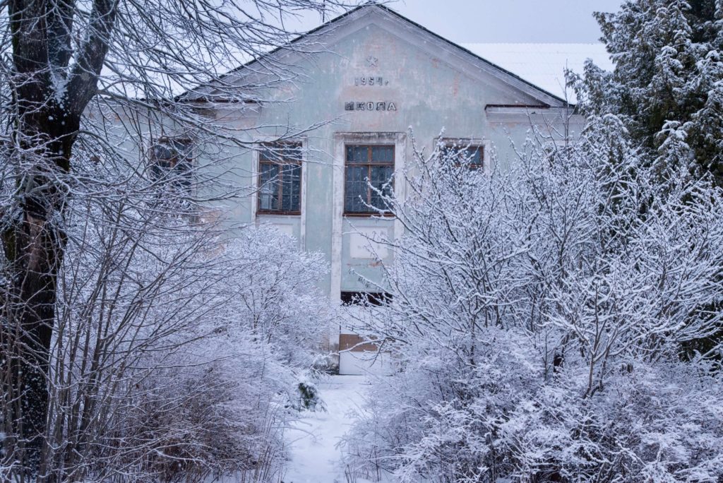 Photo of a white schoolhouse closely surrounded by snow covered trees and bushes. There are no obvious signs of abandonment. Above the central window and beneath the slanted roof are, marked in the wall, a Soviet star, the date of 1954, and a single word in cyrillic.