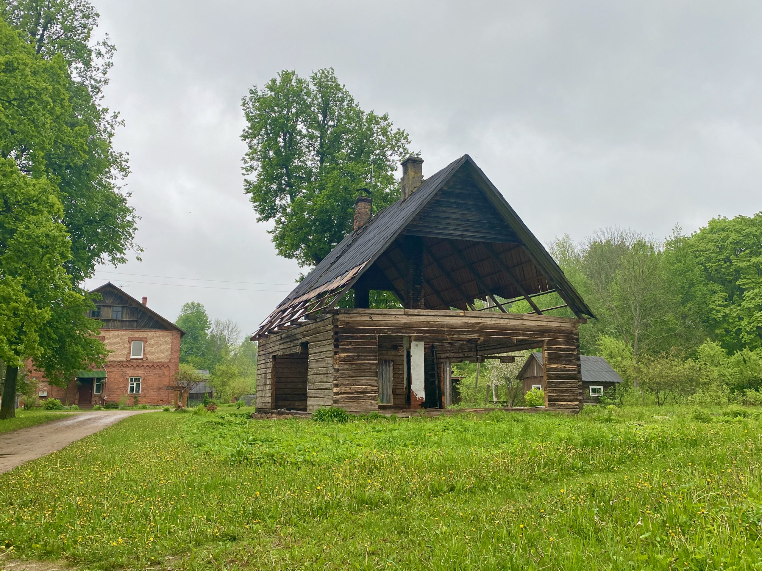 Photo of an open-roofed barn beside a large farmhouse, surrounded by bright green grass and trees.