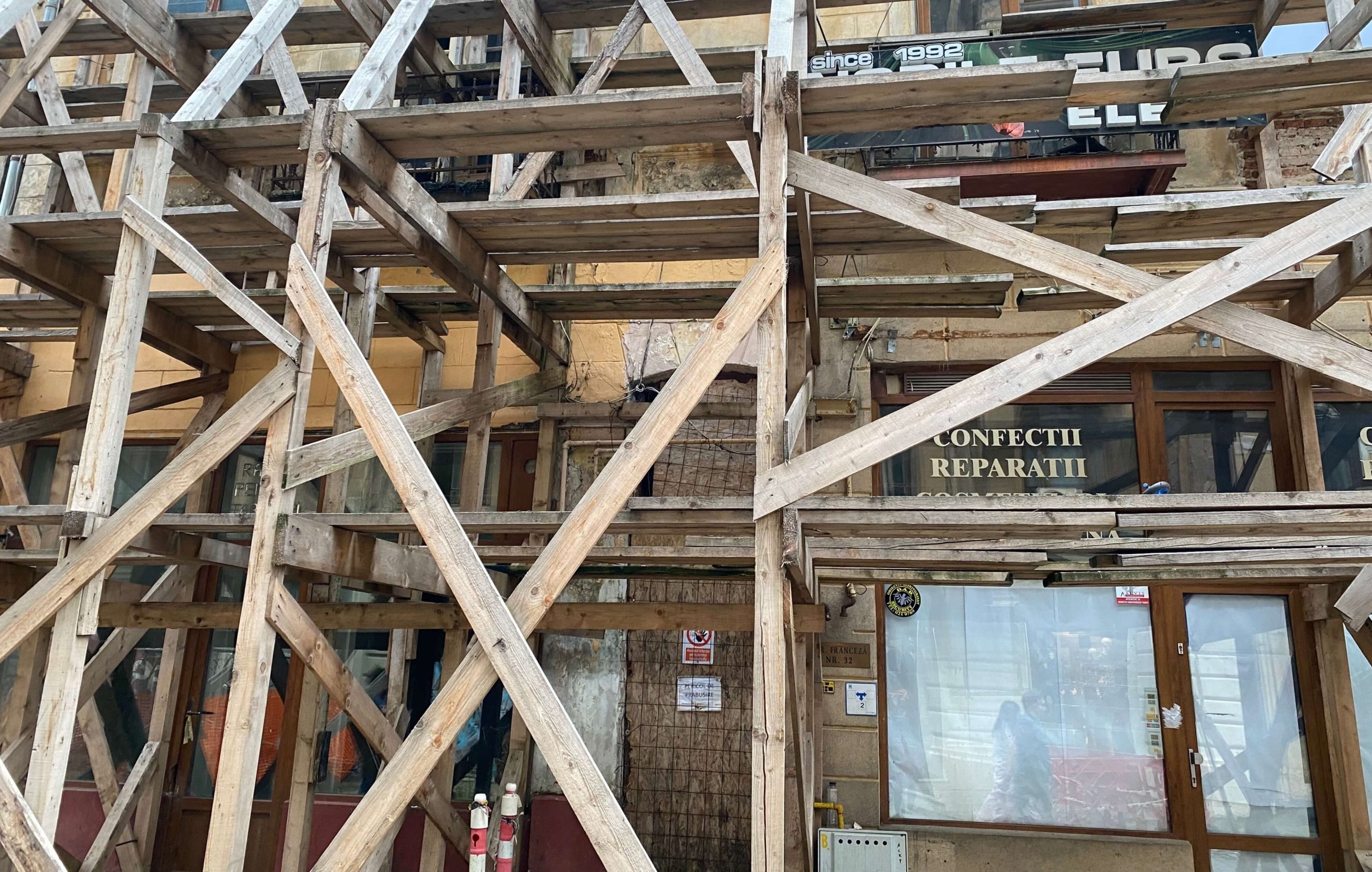Photo of a dilapidated pale stone building hidden behind a large wooden scaffold structure. Glass windows on the ground floor reflect a couple of passersby